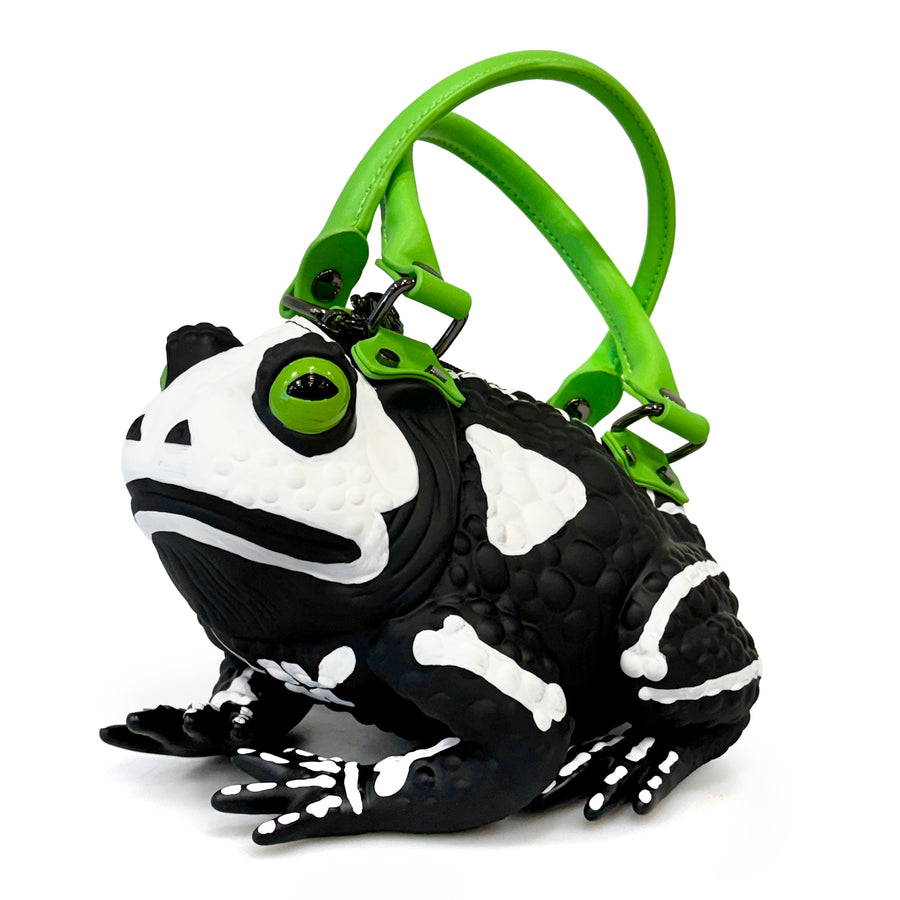 A novelty handbag shaped like a black and white skeleton toad with a bright green handle and green eyes, shown on a plain white background.